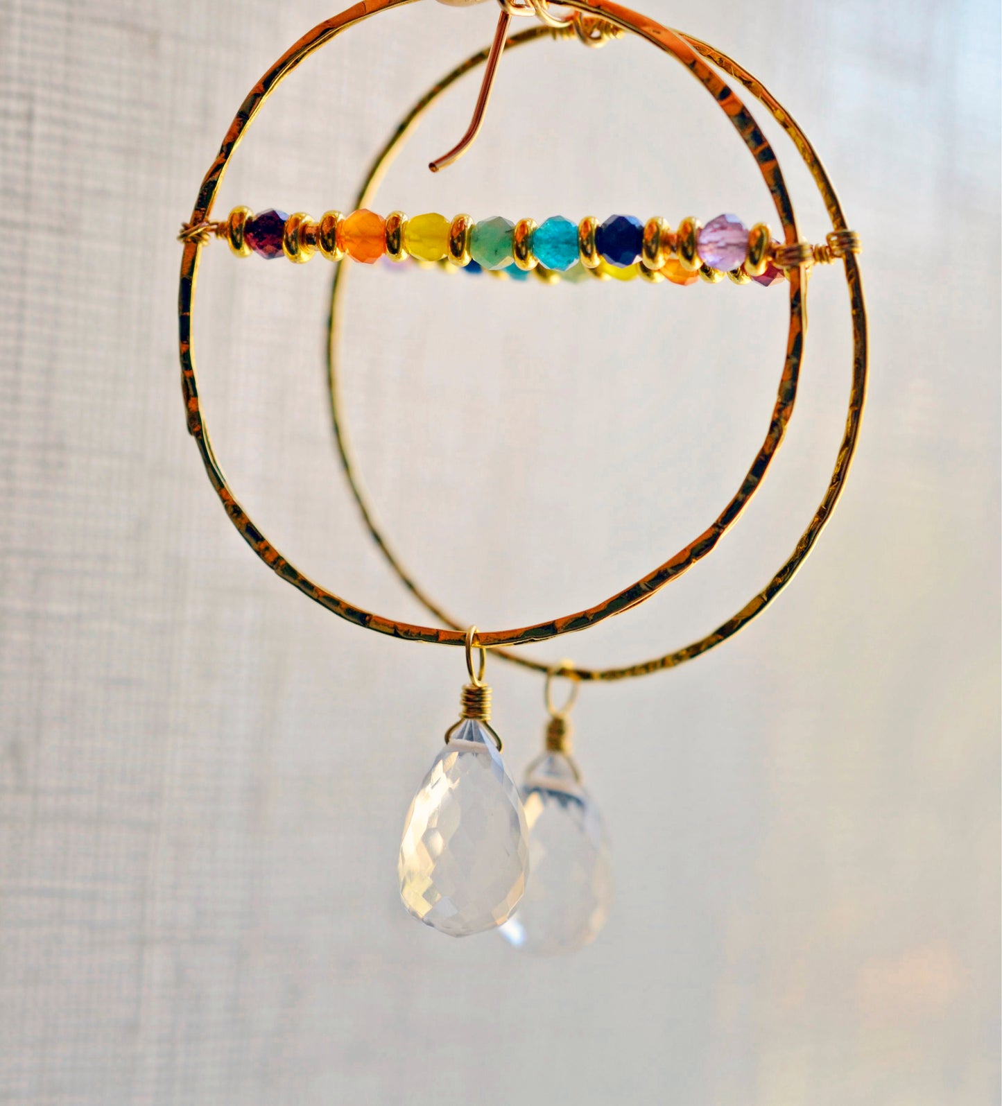 Large hammered hoop earrings with a bar of rainbow crystals spanning its center. The hoop is textured and has natural clear crystal quartz teardrop dangles. The gold style is shown.
