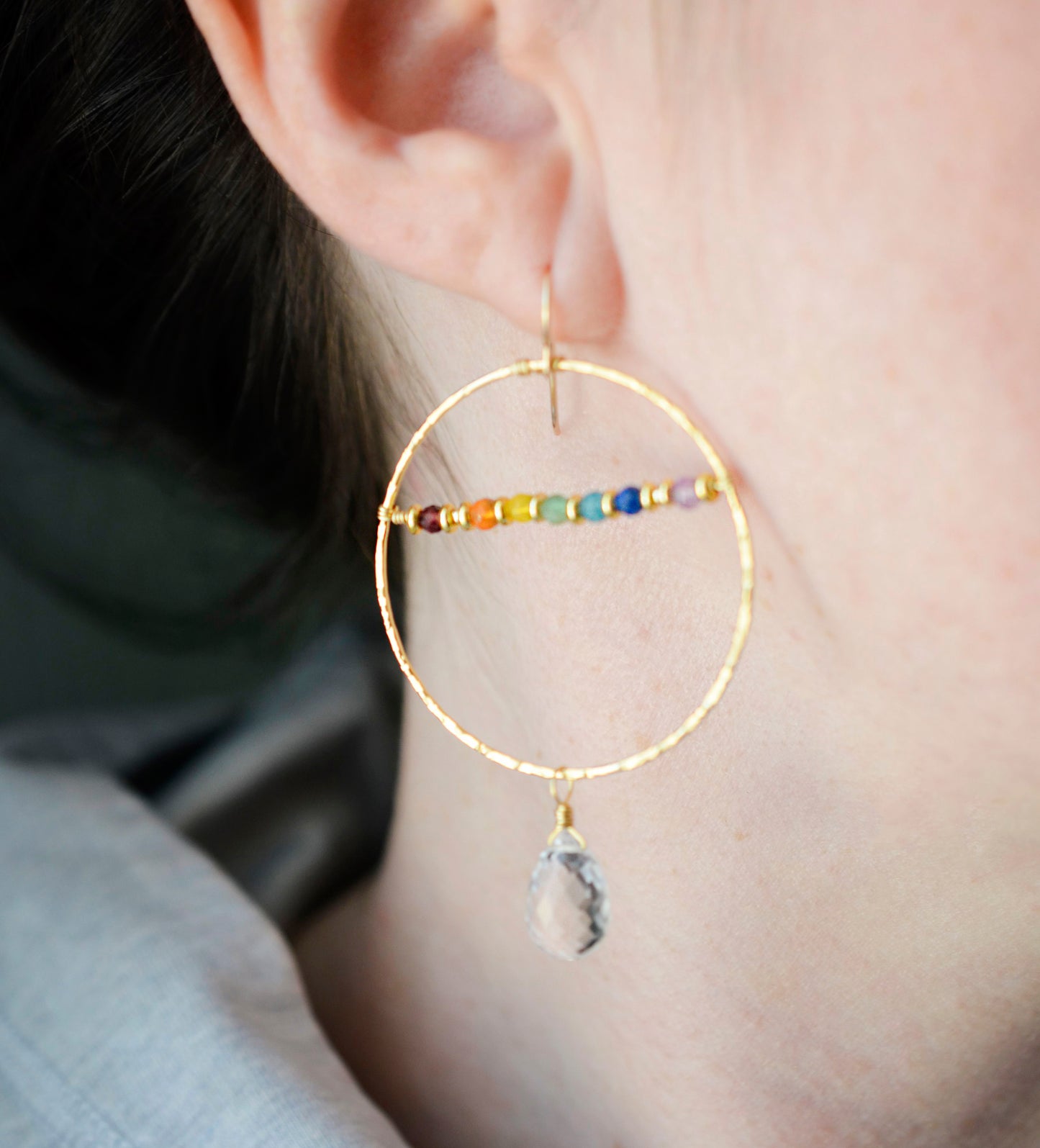 Modeled image. Large hammered hoop earrings with a bar of rainbow crystals spanning its center. The hoop is textured and has natural clear crystal quartz teardrop dangles. The gold style is shown.