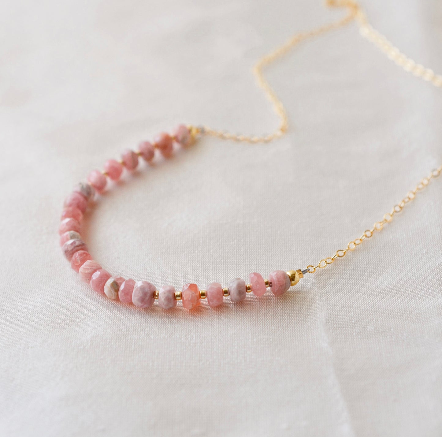 Natural pink Rhodochrosite beaded handmade necklace shown in 14k gold filled. The gemstones vary in shades of pink, peach, white, and gray and are faceted rondelle shape. They span approximately 4 inches across the front of the necklace, then attach to a dainty cable chain. Also available in sterling silver.