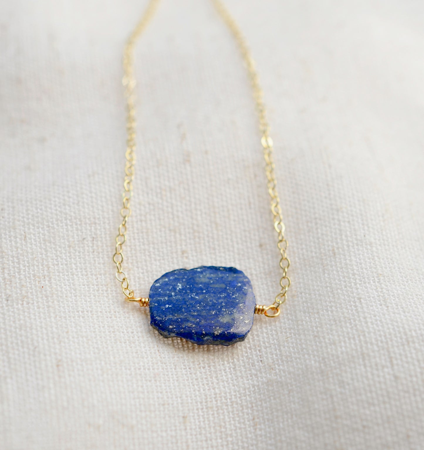 Natural blue lapis lazuli slice set onto a 14k gold filled chain. The stone is smooth polished with raw edges and oval in shape. Natural pyrite and calcite flecks are visible within each stone. 