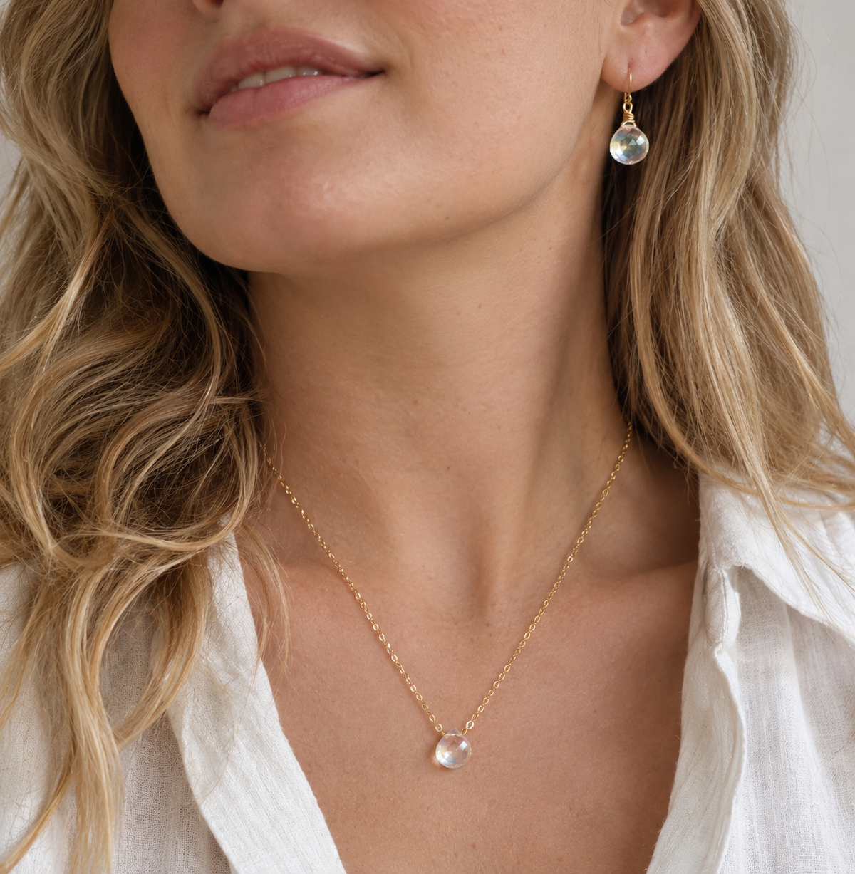 A woman wearing a mystic topaz teardrop necklace and earring set in 14k gold filled. The dainty crystals are clear but shift rainbow colors with movement. Also available in sterling silver.