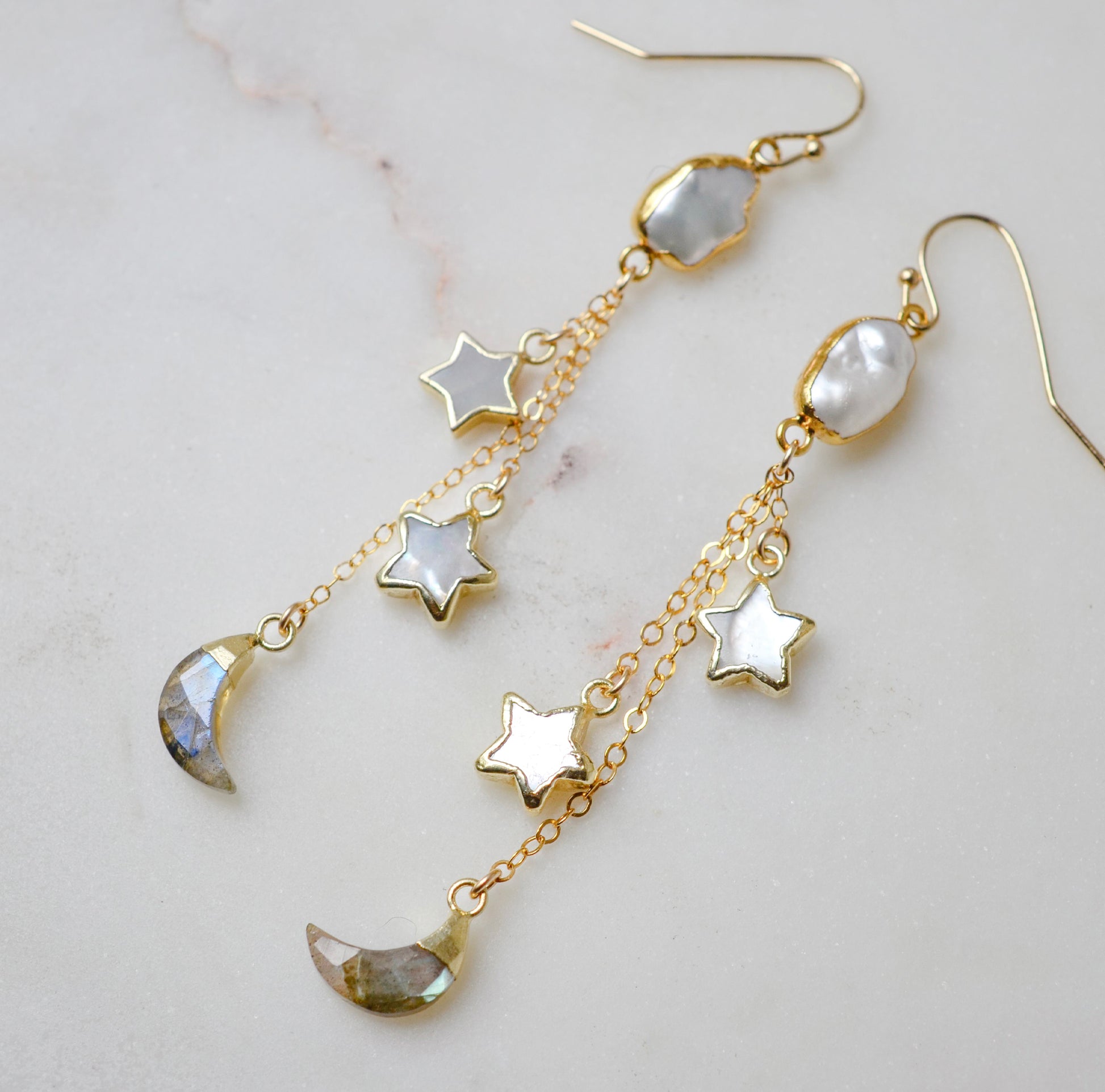Close up image. Long gold dangle, celestial earrings with star and moon charms on a light background. The stones are freshwater pearl, labradorite, and mother of pearl.