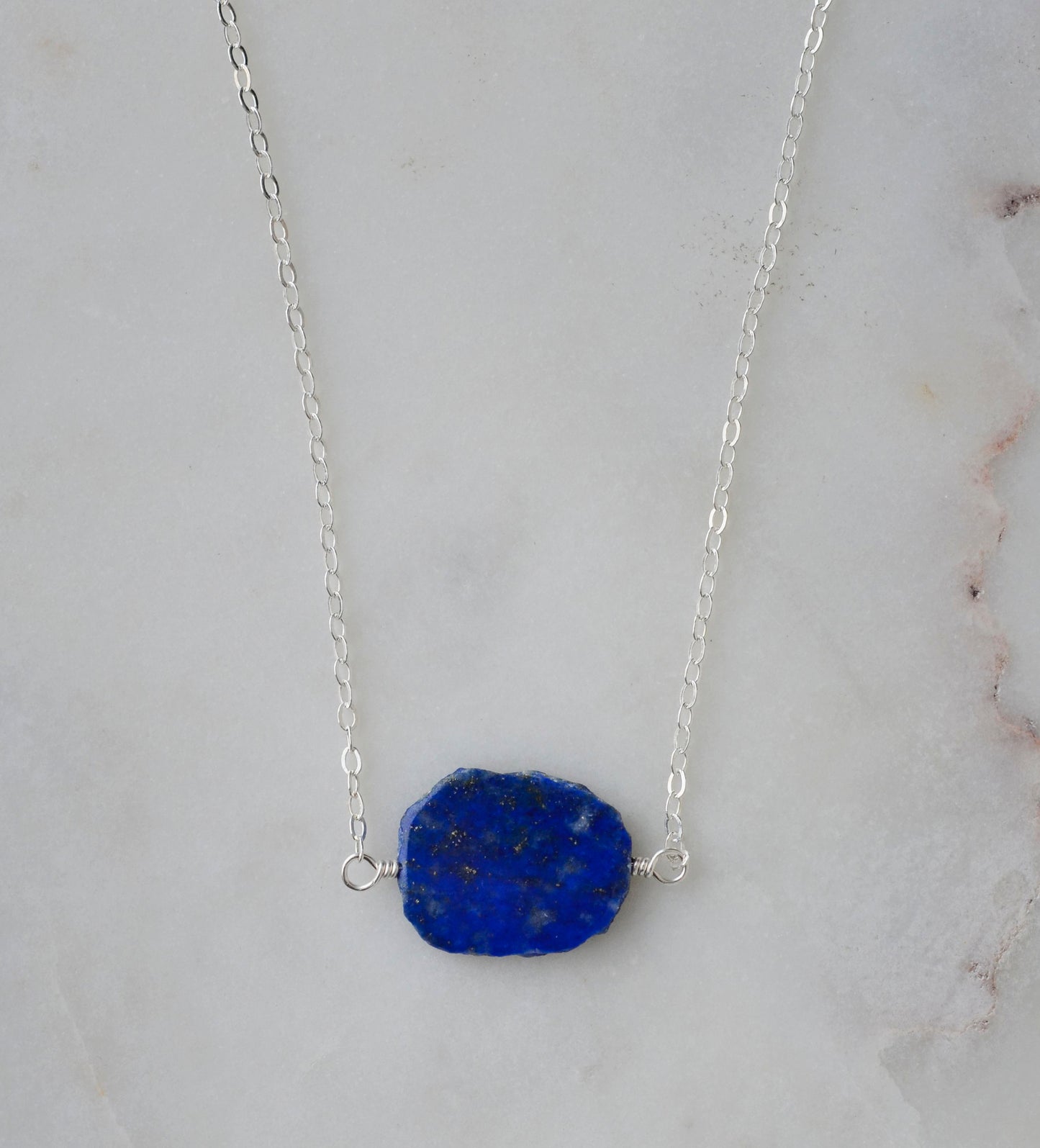 Natural blue lapis lazuli slice set onto a sterling silver chain. The stone is smooth polished with raw edges and oval in shape. Natural pyrite and calcite flecks are visible within each stone. 