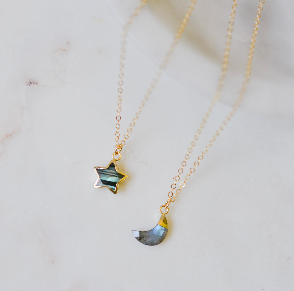Two delicate gold chain necklaces with star and crescent moon pendants featuring natural labradorite gemstones, displayed on a light marble surface. Each necklace is handmade by Gemnia in Connecticut.