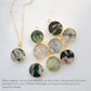 A dainty stone necklace with moss agate pendant on a light background. Shown with other stones to explain color variation. Each stone is different with some having a lot of moss, or dendritic, inclusions, while others are more clear with minimal green inclusions. This necklace is handmade in Connecticut by Gemnia Jewelry.