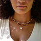 A single white cowrie shell pendant hangs from a 14k gold filled chain necklace. Modeled with multiple beaded gemstone necklaces.