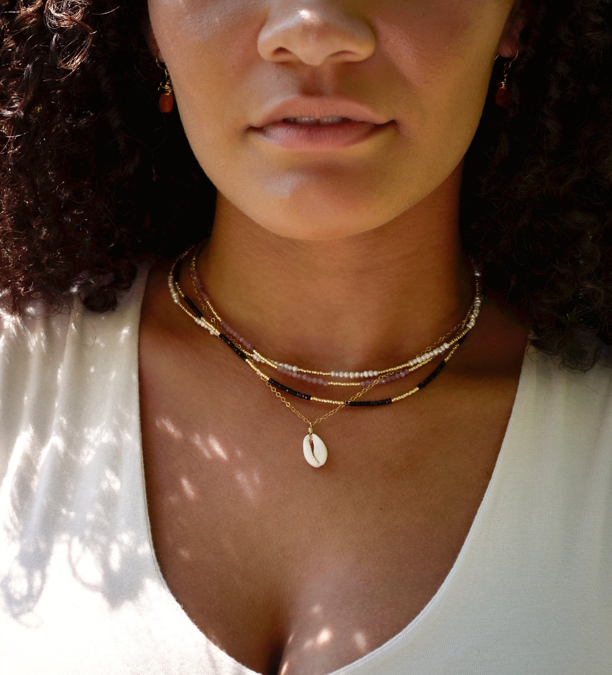 A single white cowrie shell pendant hangs from a 14k gold filled chain necklace. Modeled with multiple beaded gemstone necklaces.