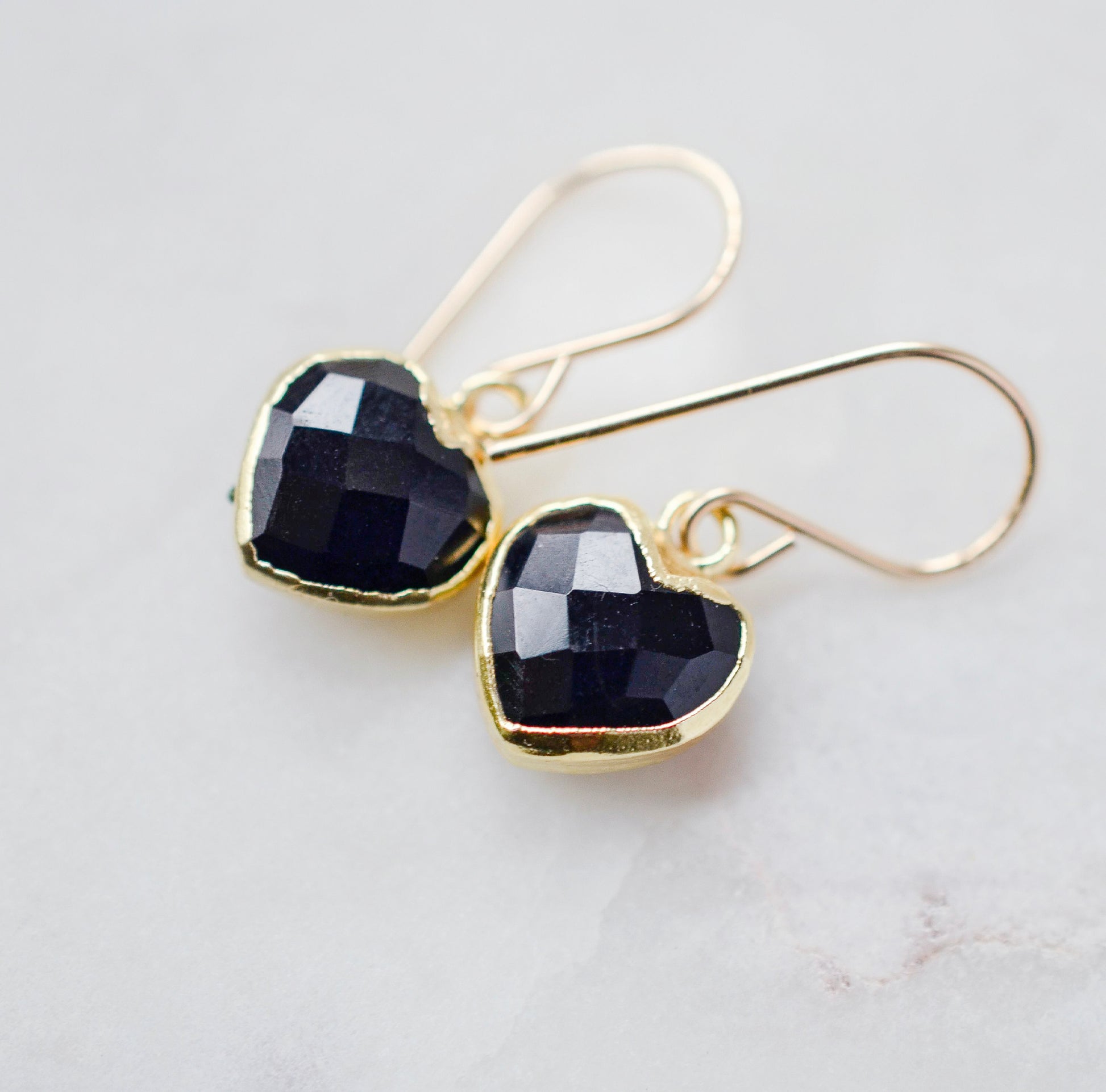 Black onyx heart earrings in gold. The stones are faceted and solid black with gold bezels surrounding their sides. The ear wires are basic hooks.