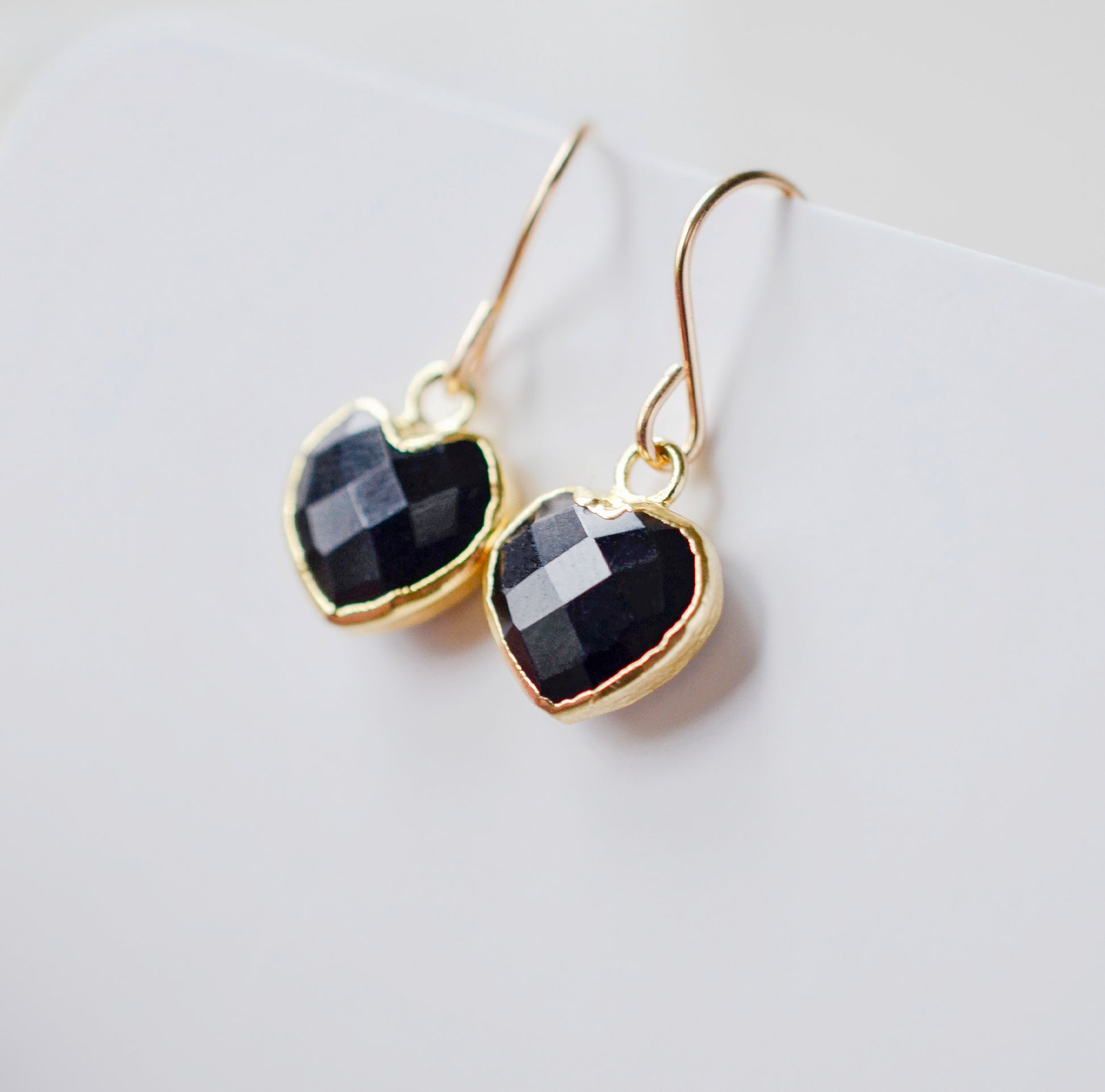 Black onyx heart earrings in gold. The stones are faceted and solid black with gold bezels surrounding their sides. The ear wires are basic hooks.