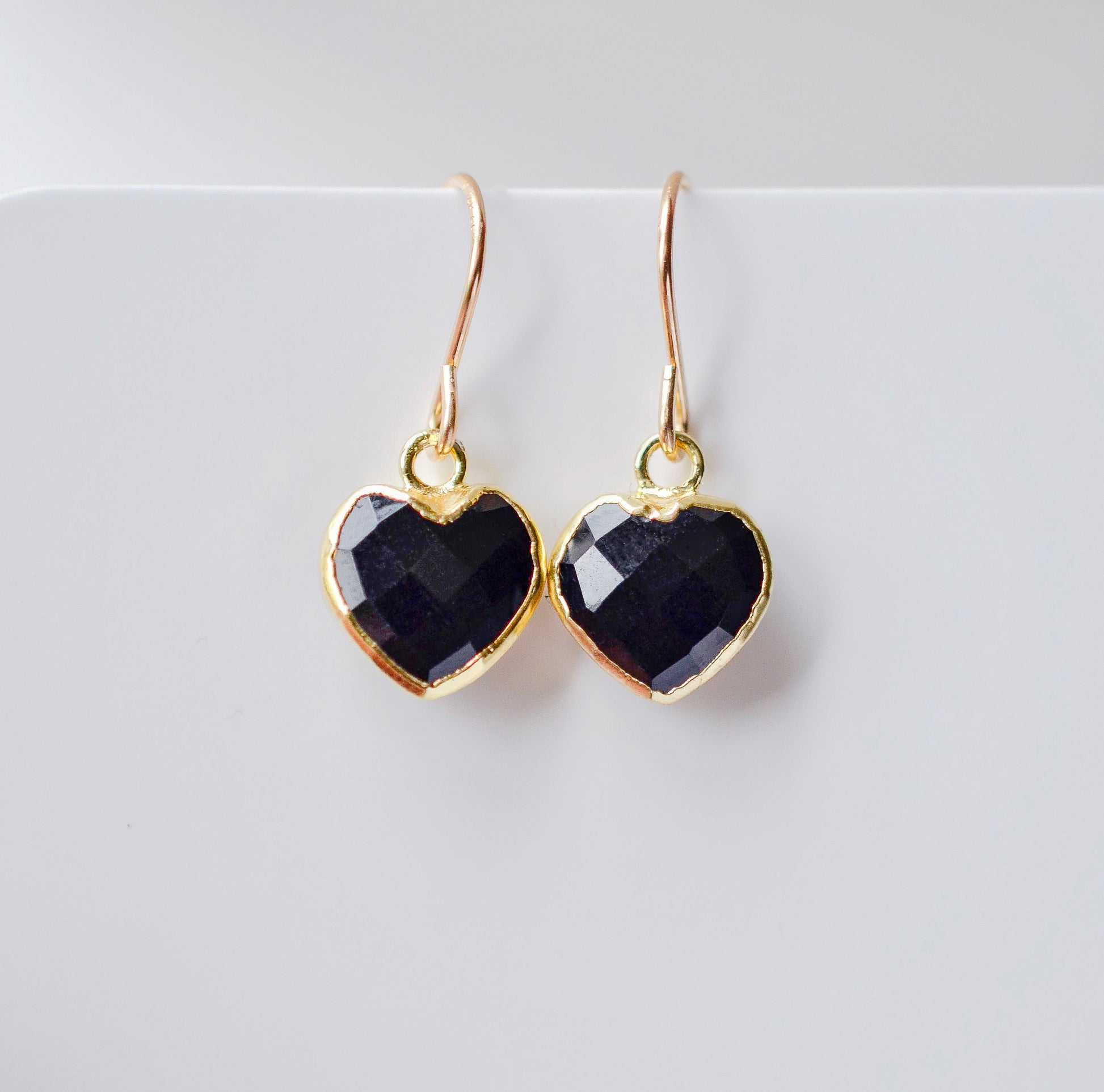 Black onyx heart earrings in gold. The stones are faceted and solid black with gold bezels surrounding their sides. The ear wires are basic hooks.