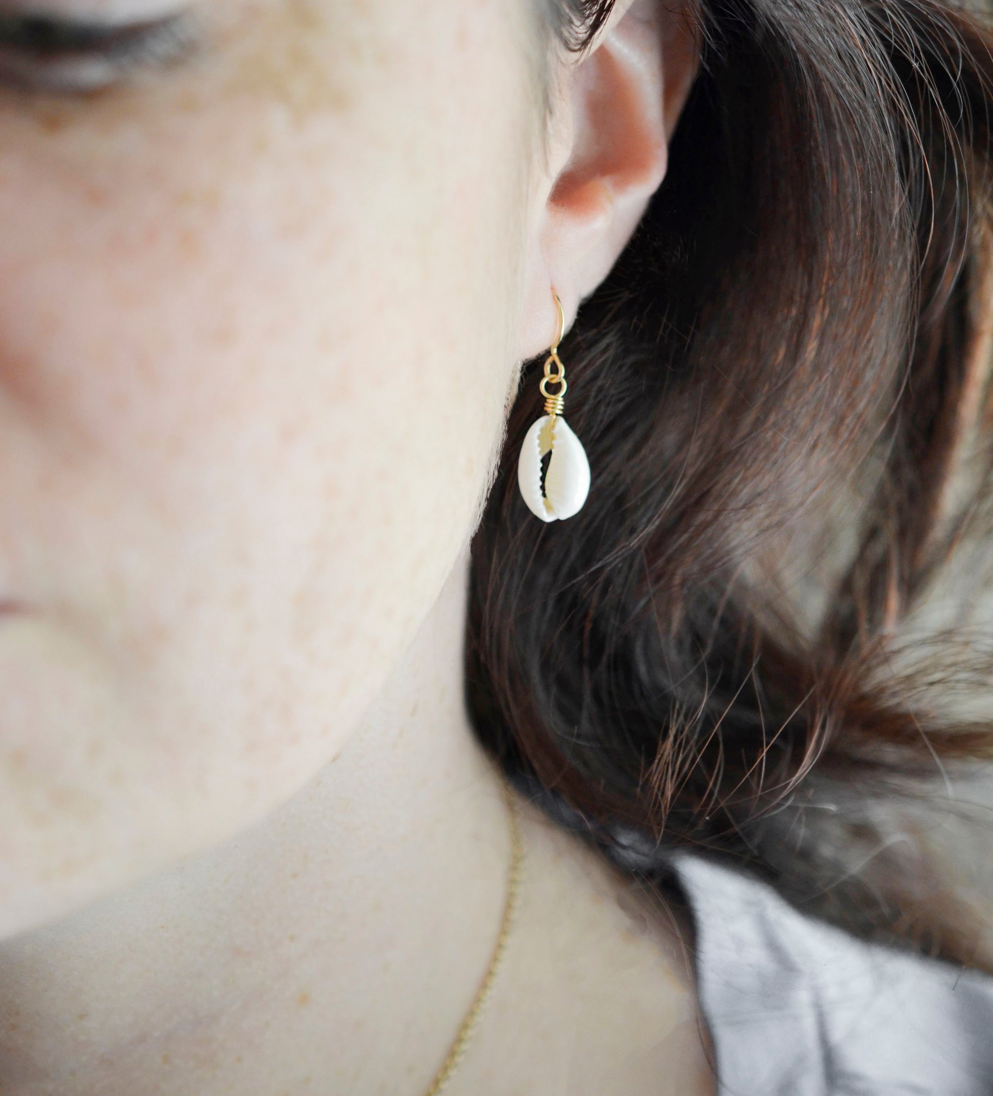 A natural off-white cowrie shell hangs from an earring hook. Shown in gold. Modeled image.