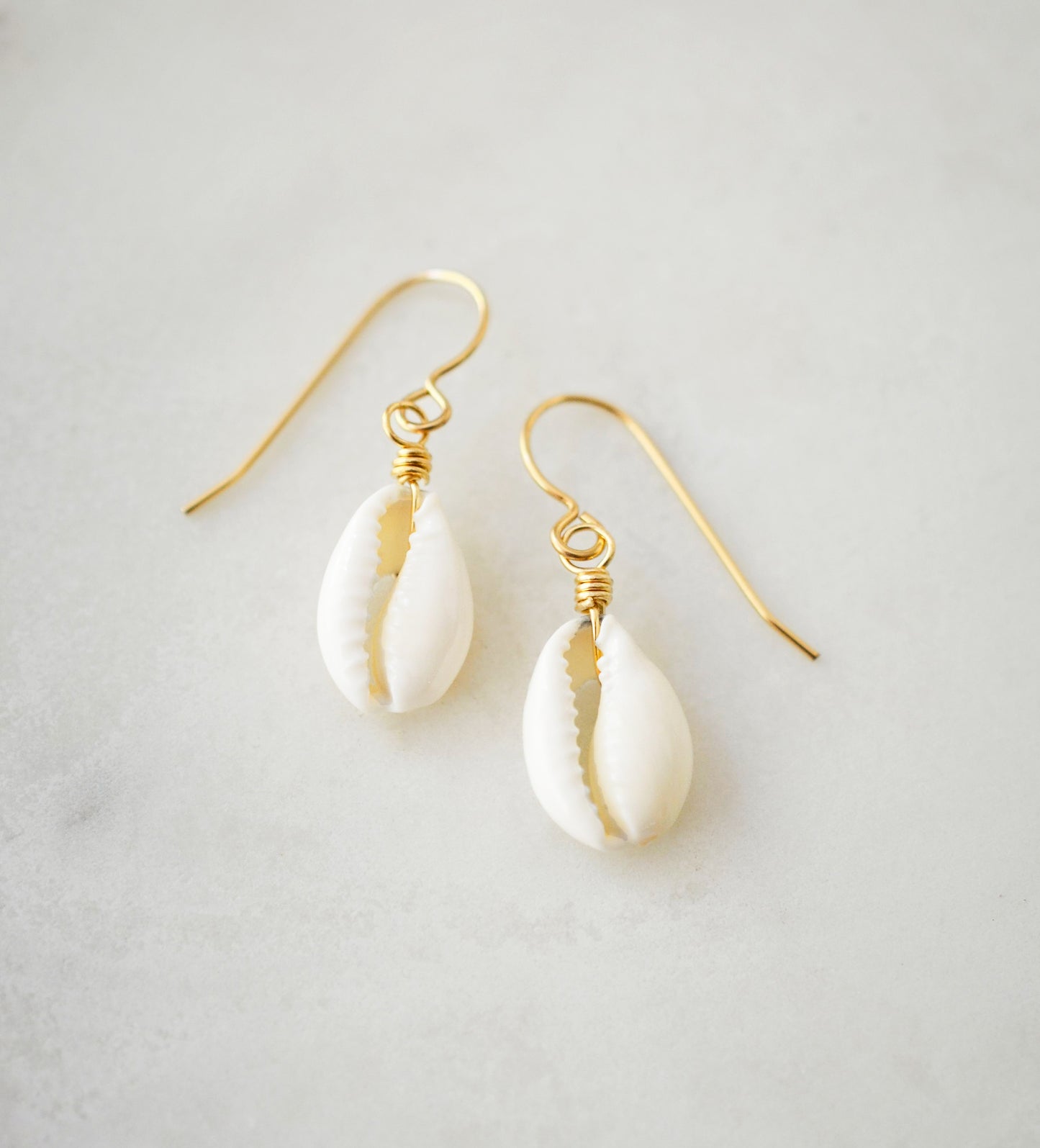 A natural off-white cowrie shell hangs from an earring hook. Shown in gold.