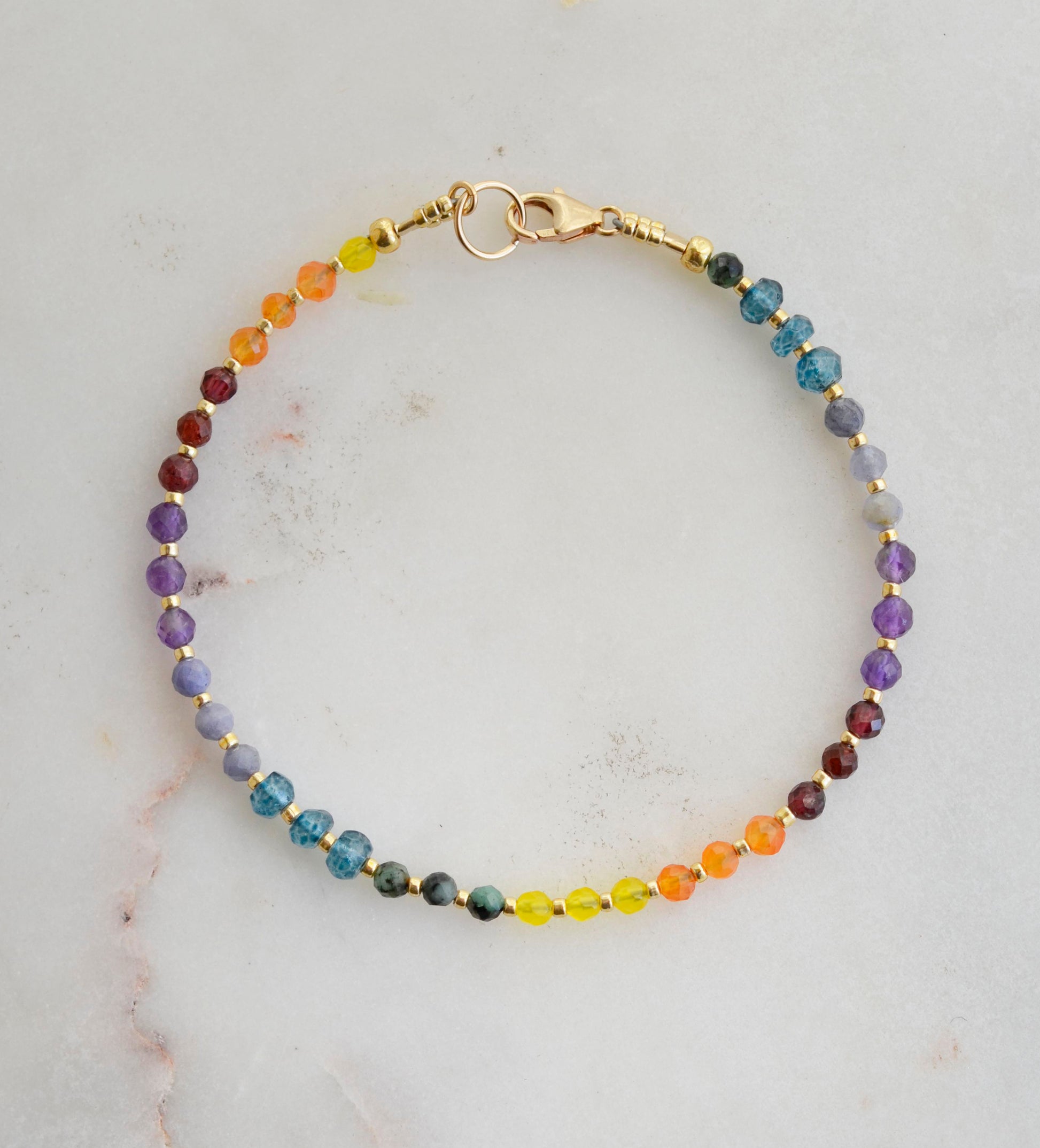 Beaded gemstone bracelet in rainbow colors. Handmade with natural gemstones including: yellow agate, orange carnelian, wine-red garnet, amethyst, tanzanite, topaz, and green emerald. Shown with a gold clasp.