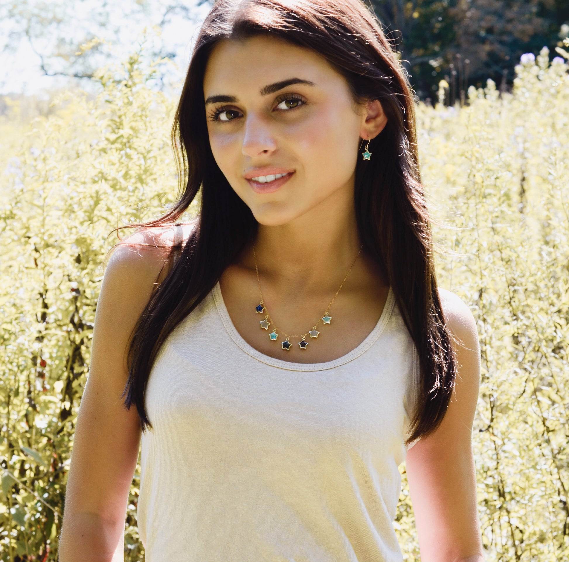 Woman wearing a dainty necklace with star-shaped pendants on a 14k gold filled chain. Each star is a different gemstone. Stones include: Lapis lazuli, rainbow moonstone, turquoise, labradorite, amethyst, rose quartz, and amazonite. This jewelry is handmade in New England by Gemnia.