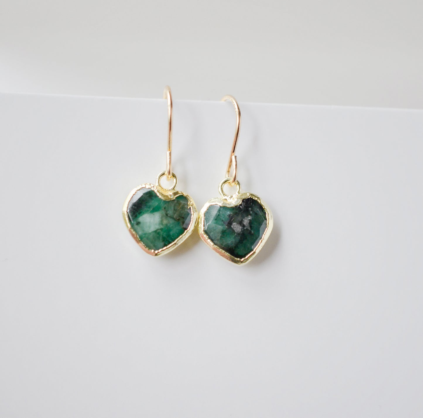 Raw emerald heart earrings in gold. The stones are bezeled in 22k gold then set on gold earwires. The gemstone colors range in dark greens with hints of white or black.