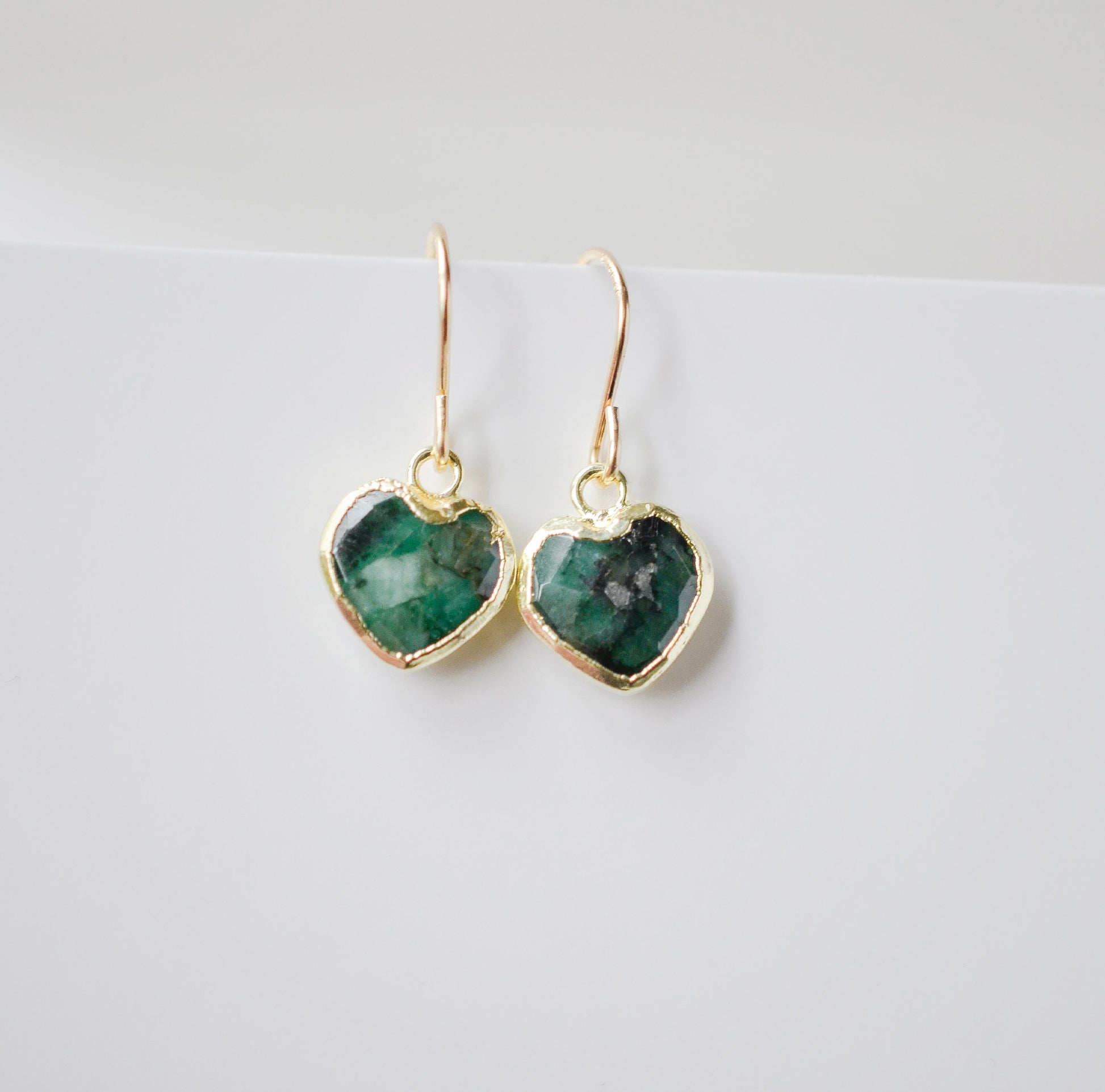 Raw emerald heart earrings in gold. The stones are bezeled in 22k gold then set on gold earwires. The gemstone colors range in dark greens with hints of white or black.