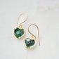 Raw emerald heart earrings in gold. The stones are bezeled in 22k gold then set on gold earwires. The gemstone colors range in dark greens with hints of white or black.