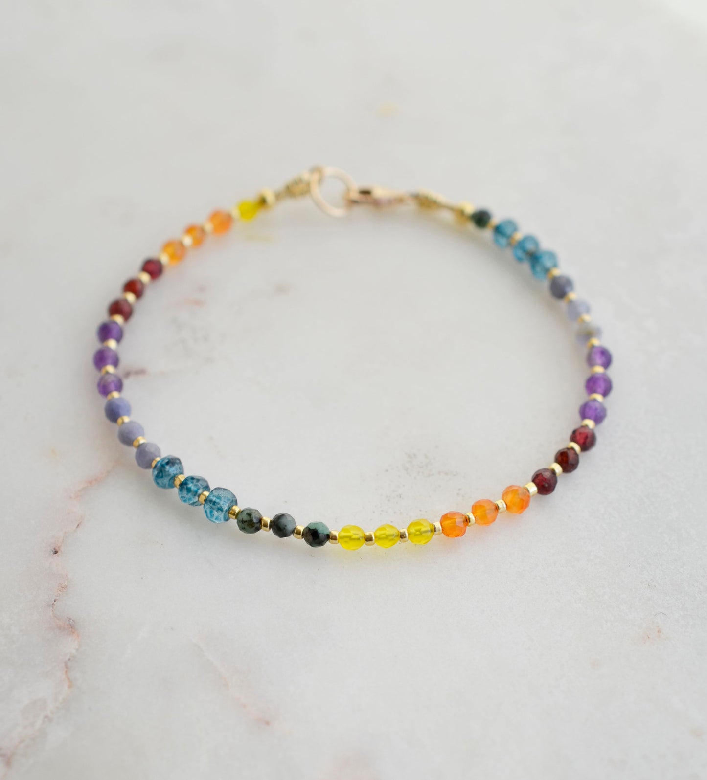 Beaded gemstone bracelet in rainbow colors. Handmade with natural gemstones including: yellow agate, orange carnelian, wine-red garnet, amethyst, tanzanite, topaz, and green emerald. Shown with a gold clasp.