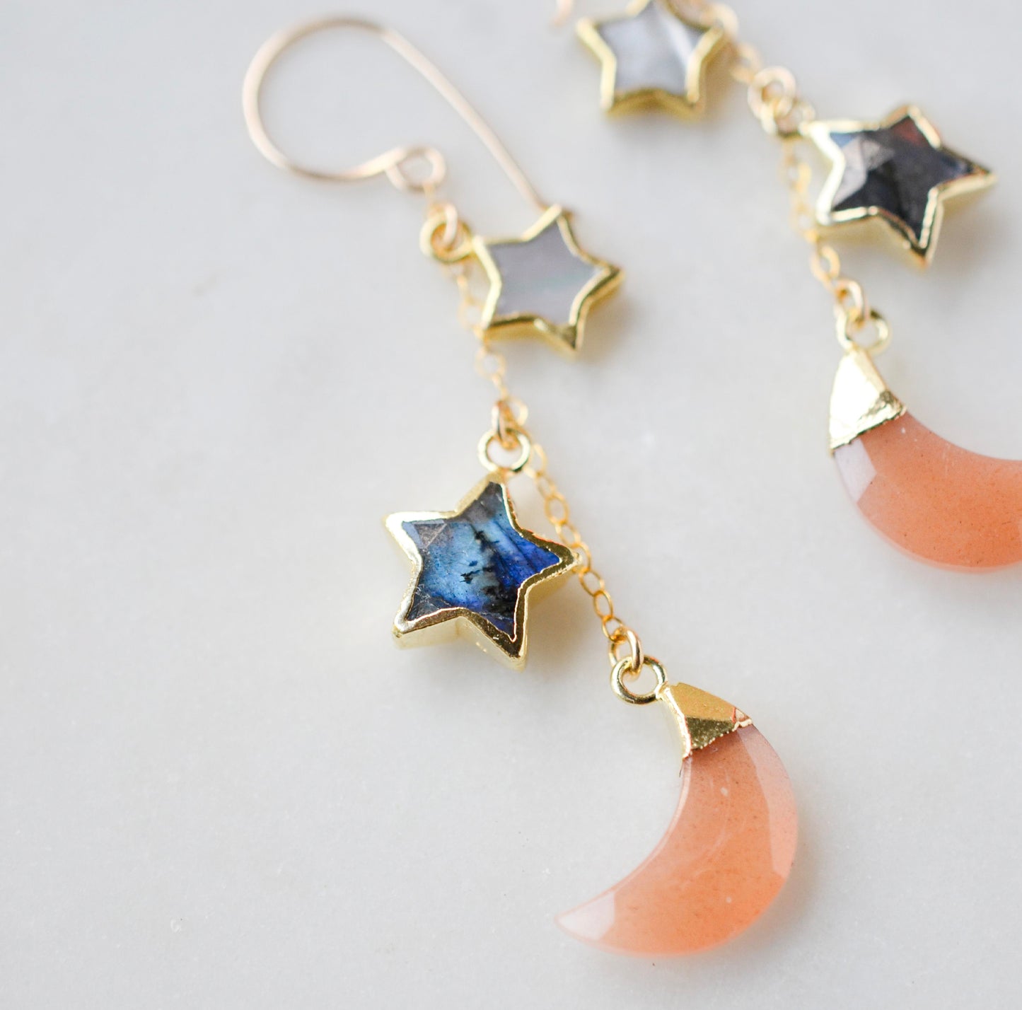 Mother of pearl, Labradorite, and Peach Moonstone moon and star dangle earrings in gold. Each earring has two stars hanging over a peach moonstone crescent. Close up.