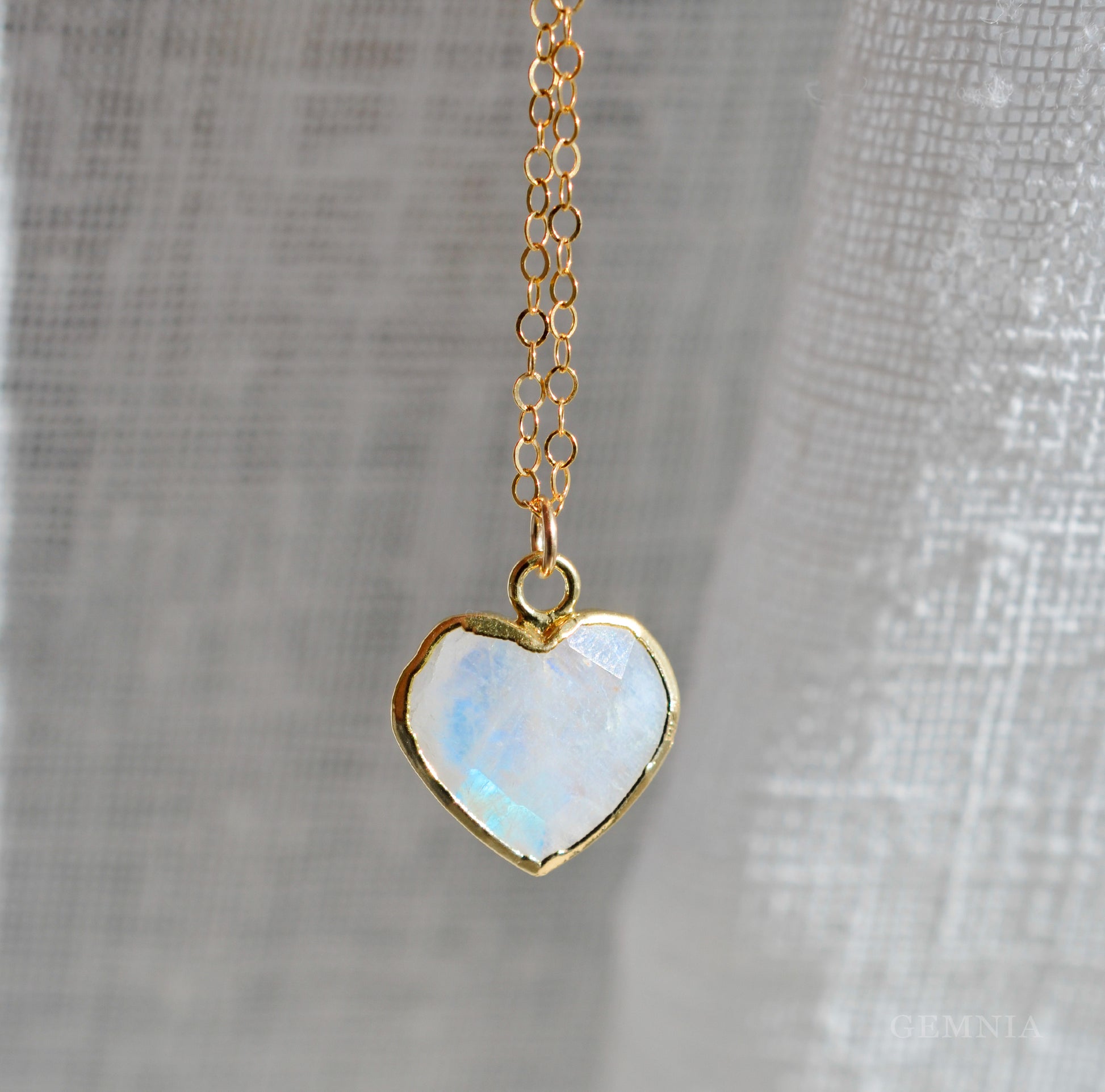 Heart-shaped rainbow moonstone pendant on a 14k gold filled chain against a gray fabric background. This dainty romantic necklace is handmade by Gemnia Jewelry in Connecticut.