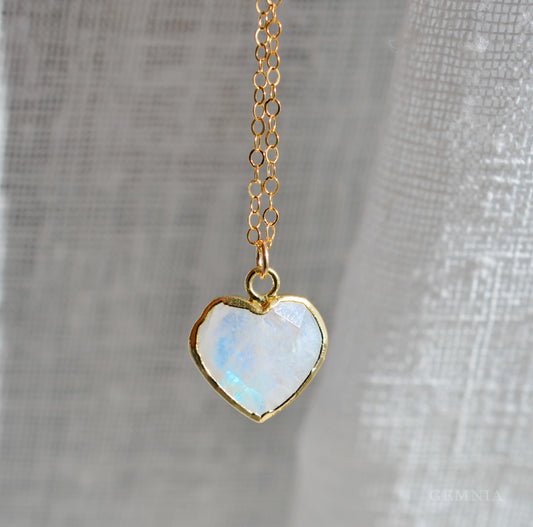 Heart-shaped rainbow moonstone pendant on a 14k gold filled chain against a gray fabric background. This dainty romantic necklace is handmade by Gemnia Jewelry in Connecticut.