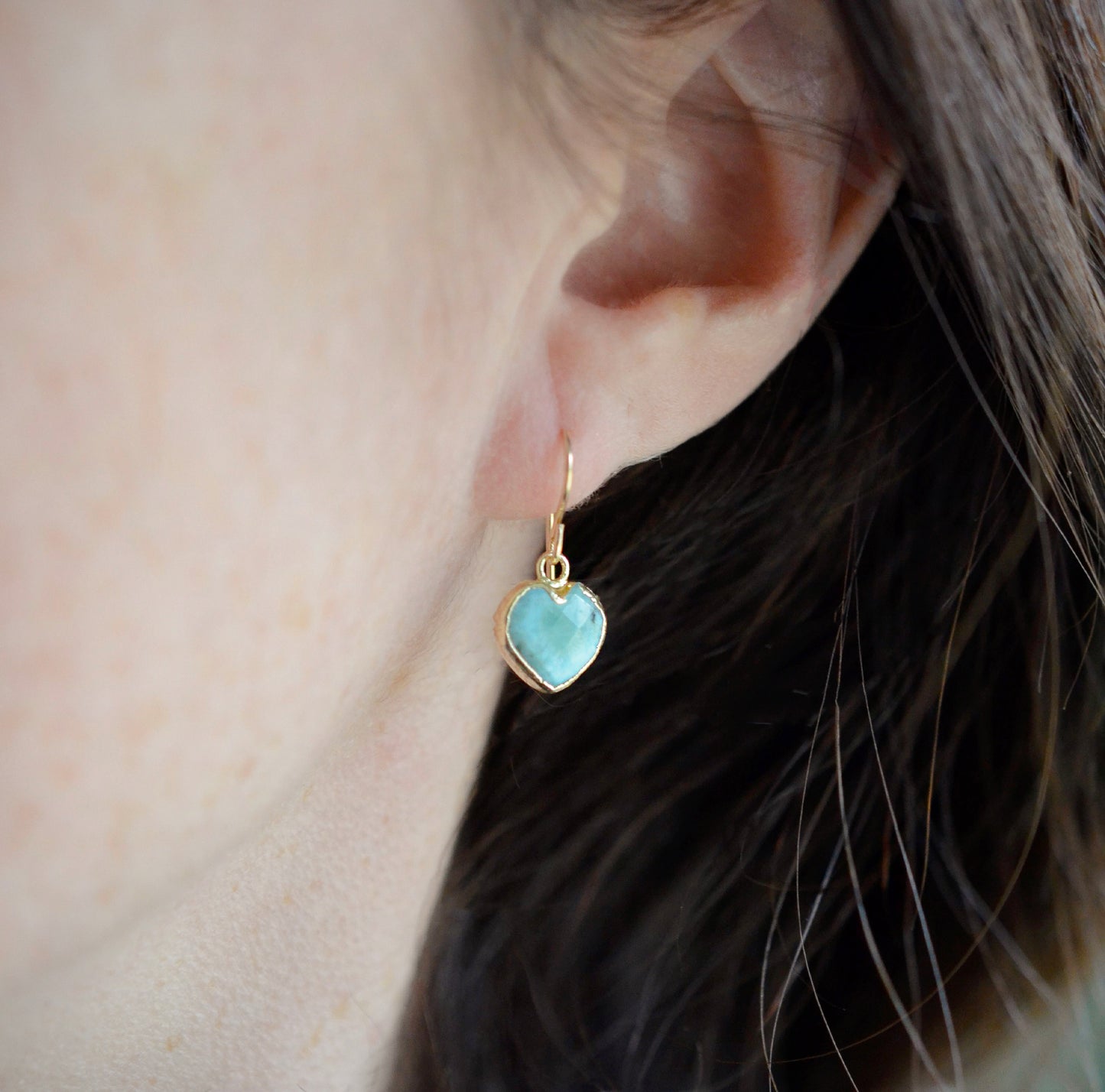 Aqua blue, natural Larimar stone heart earrings in gold. The stones range in blue, white, or green with the occasion mark of brown or black. Modeled image.