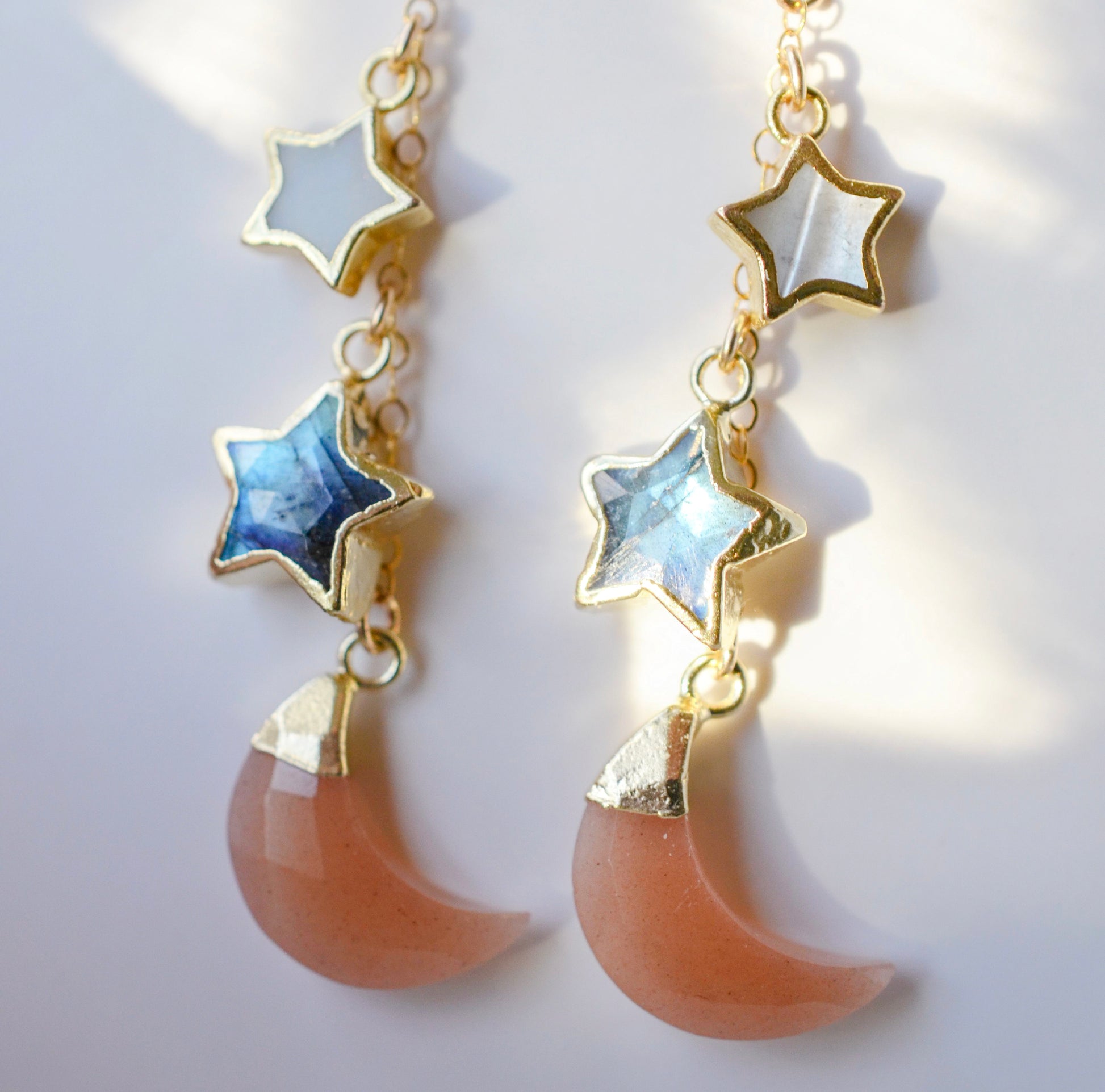 Mother of pearl, Labradorite, and Peach Moonstone moon and star dangle earrings in gold. Each earring has two stars hanging over a peach moonstone crescent. Close up image.