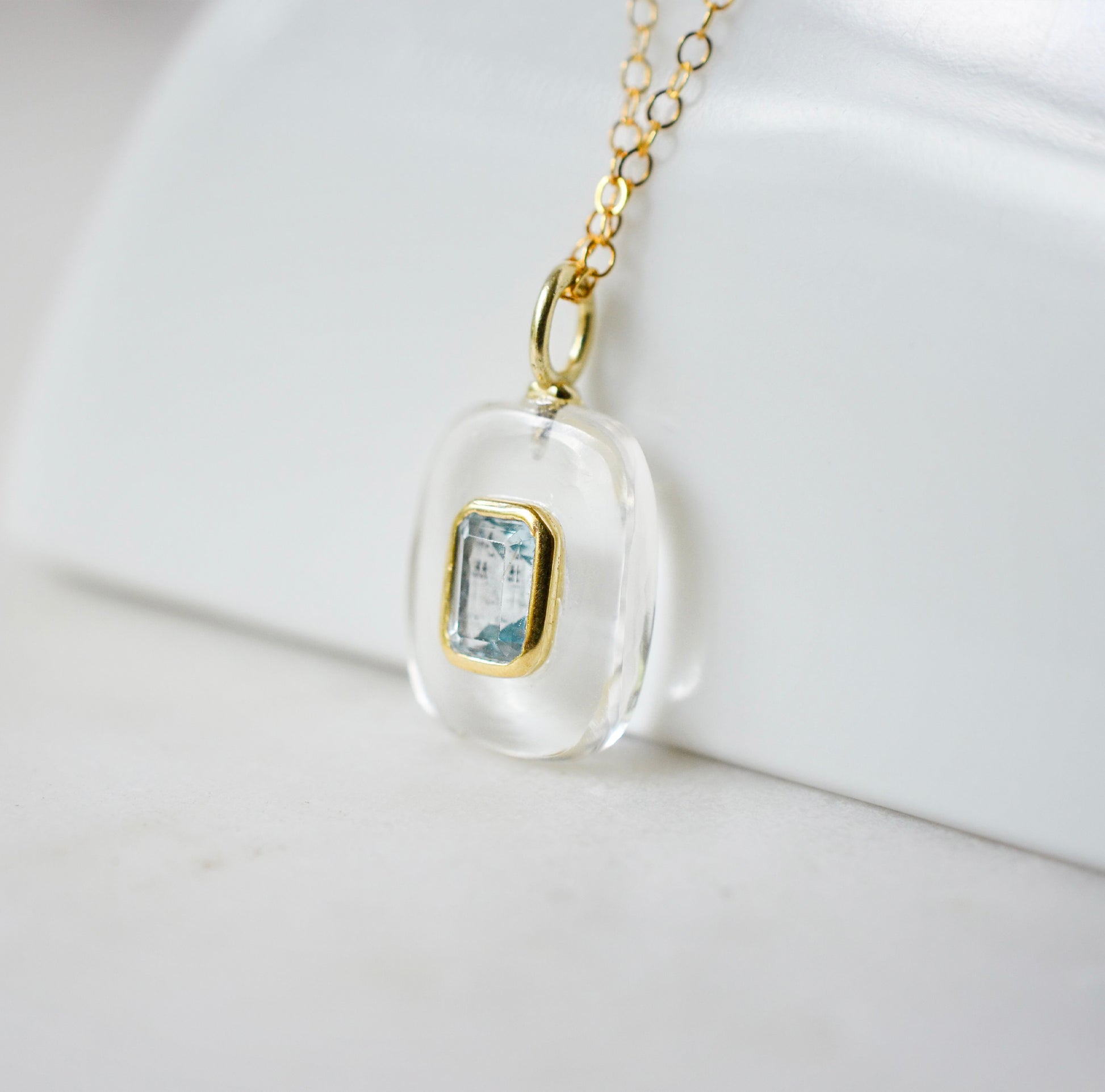 Side view. Handmade Maris necklace featuring a Blue Topaz set inside a clear Rock Crystal frame on a 14K Gold Filled chain. Ideal gift for wife, girlfriend, or mom. Minimalist topaz pendant for layering.
