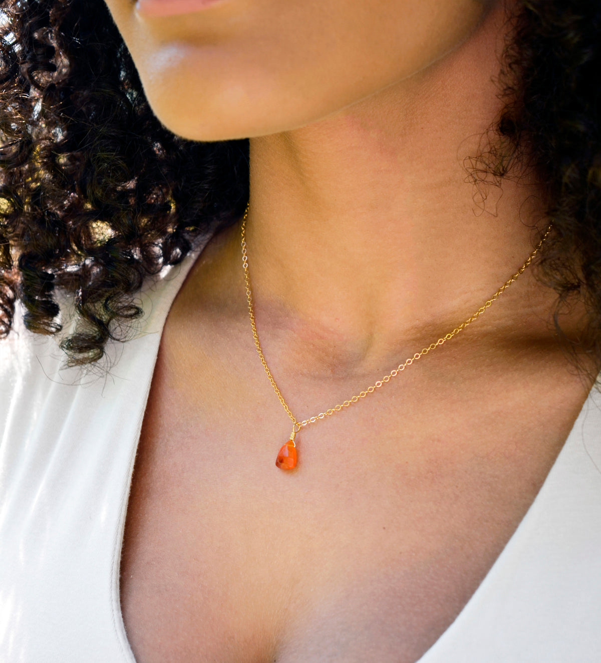 A woman wearing a natural, faceted orange Carnelian gemstone necklace with a 14k gold filled chain. The crystal is a triangular teardrop shape and ranges in shades of orange and sometimes red. Also available in sterling silver. Handmade in Connecticut by Gemnia.