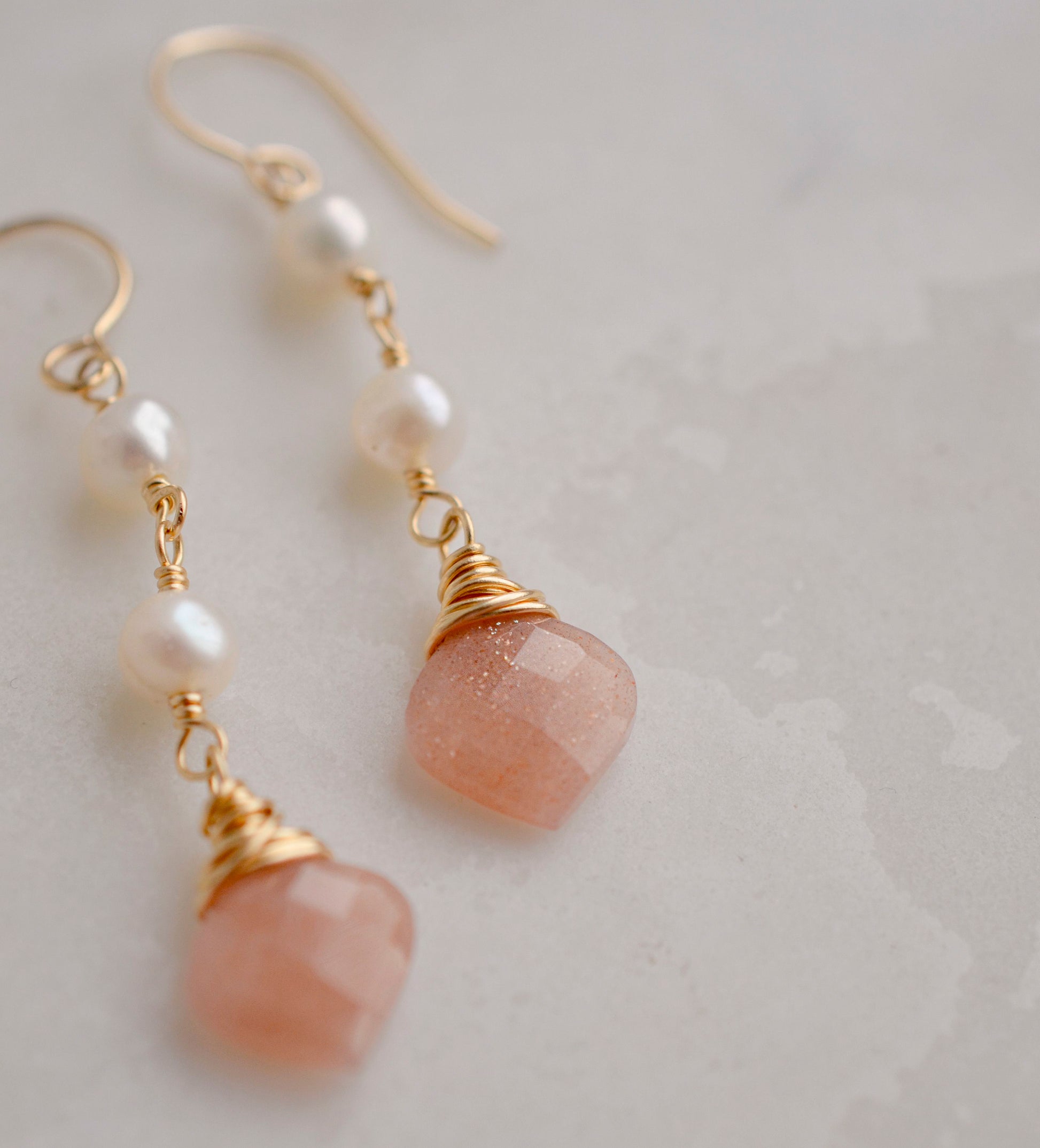 Two white semi-round pearls hang over natural peach Moonstone faceted drops. The gold style is shown.