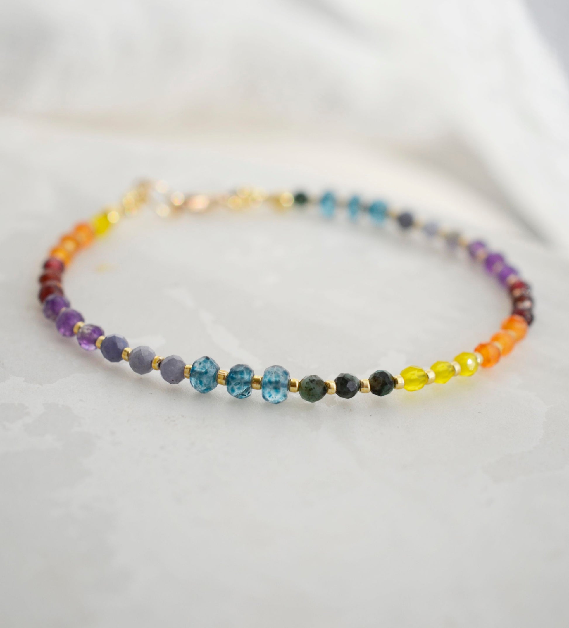 Beaded gemstone bracelet in rainbow colors. Handmade with natural gemstones including: yellow agate, orange carnelian, wine-red garnet, amethyst, tanzanite, topaz, and green emerald. Shown with a gold clasp.