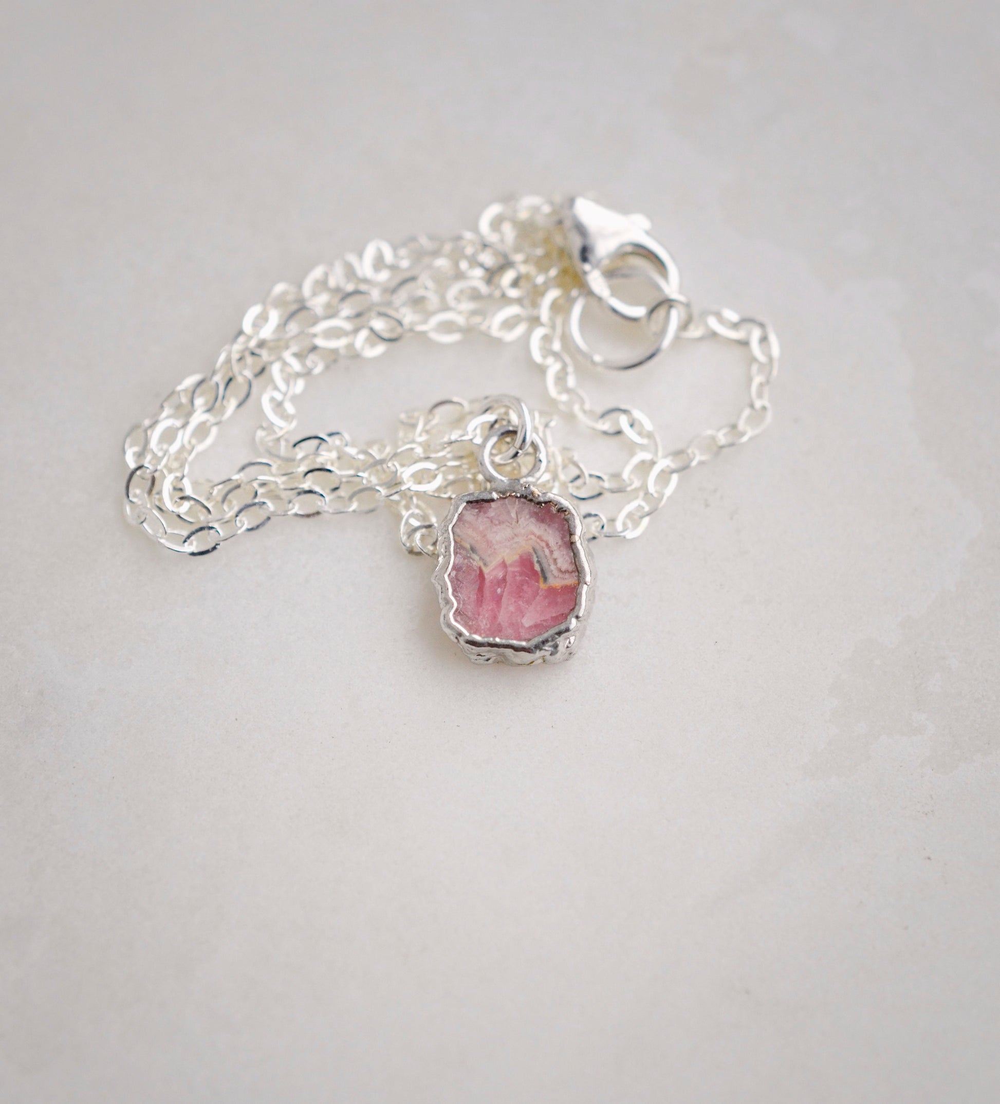 Natural pink Rhodochrosite sliced gemstone bezeled in sterling silver and set on a sterling chain. Showing a lobster claw clasp.