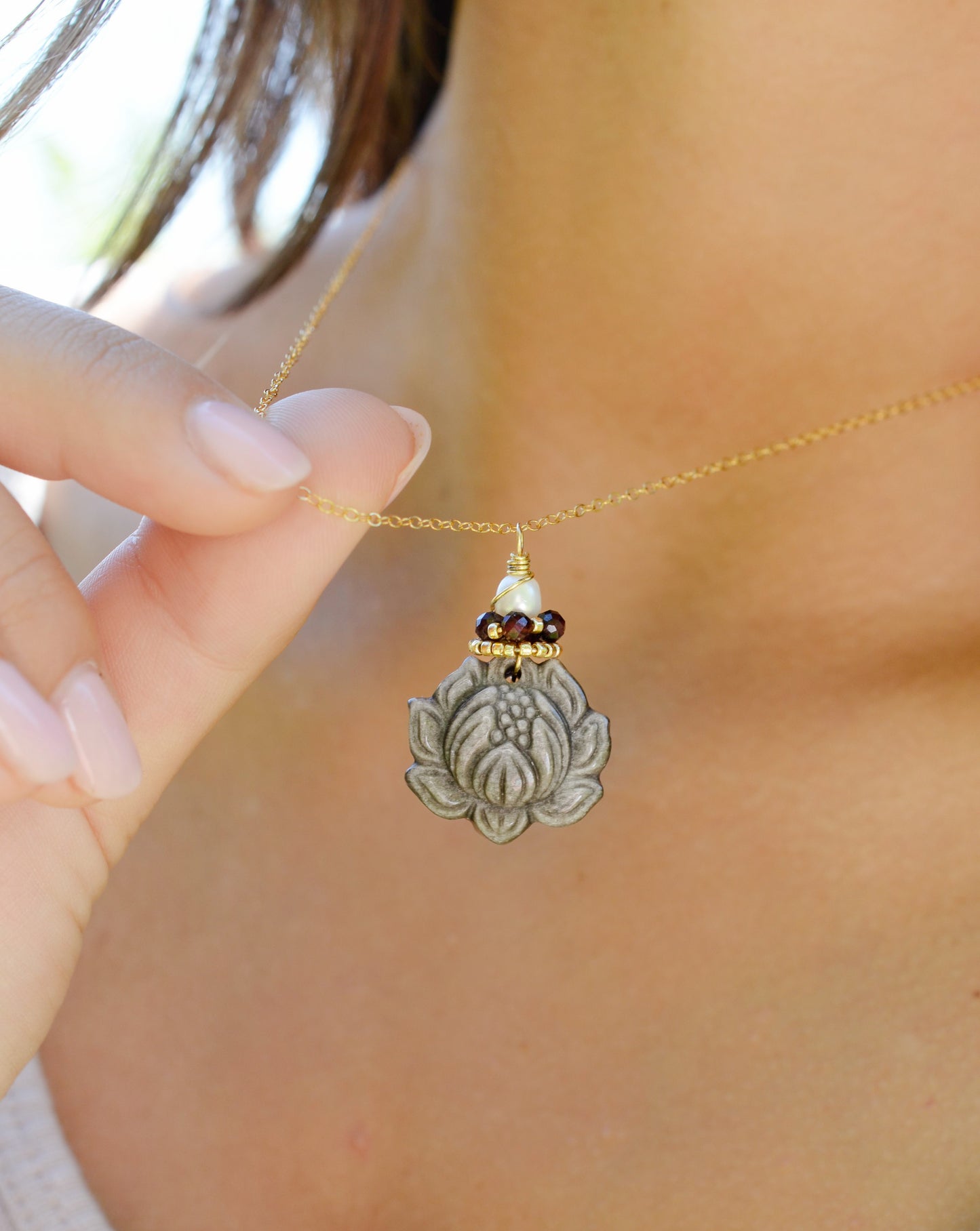 A handmade gemstone pendant featuring a carved obsidian lotus. Garnet, pearl, and gold beads decorate its top. The chain is 14k gold filled.