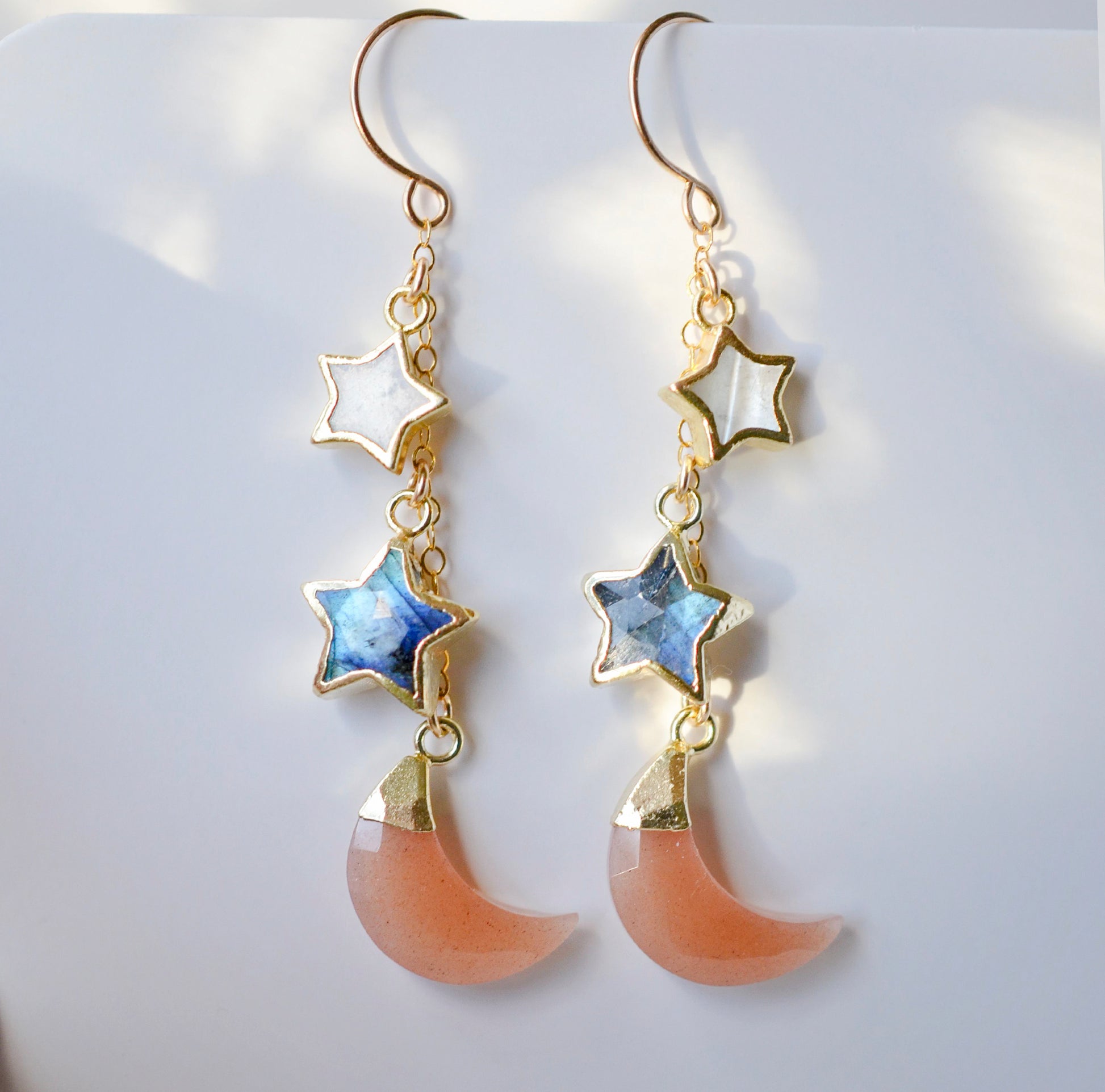 Mother of pearl, Labradorite, and Peach Moonstone moon and star dangle earrings in gold. Each earring has two stars hanging over a peach moonstone crescent.