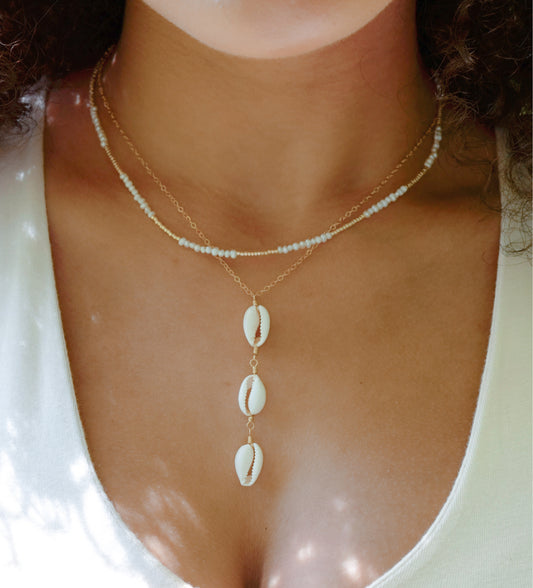 Three white cowrie shells hang from a 14k gold filled chain. Modeled with a beaded pearl necklace.
