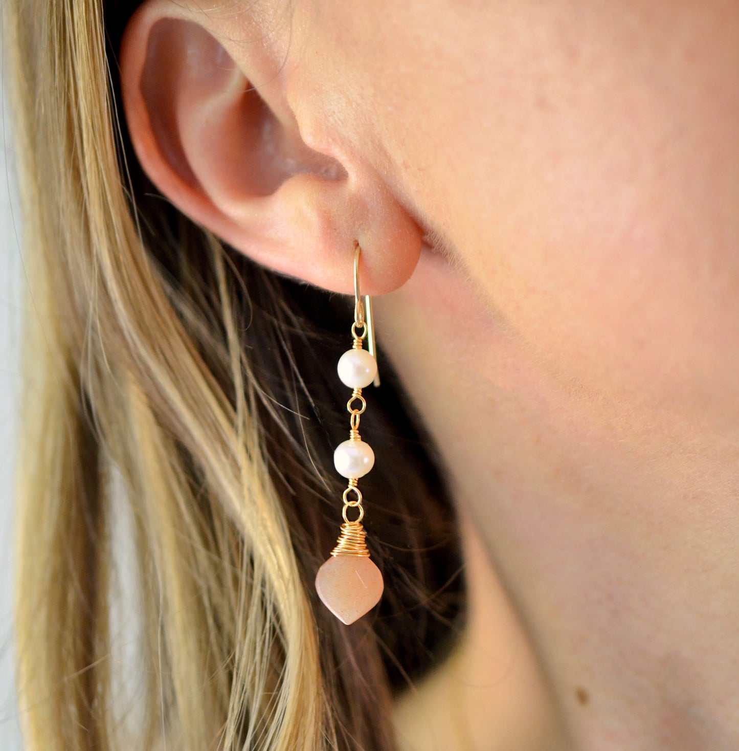 Two white semi-round pearls hang over natural peach Moonstone faceted drops. The gold style is shown. Modeled Image.