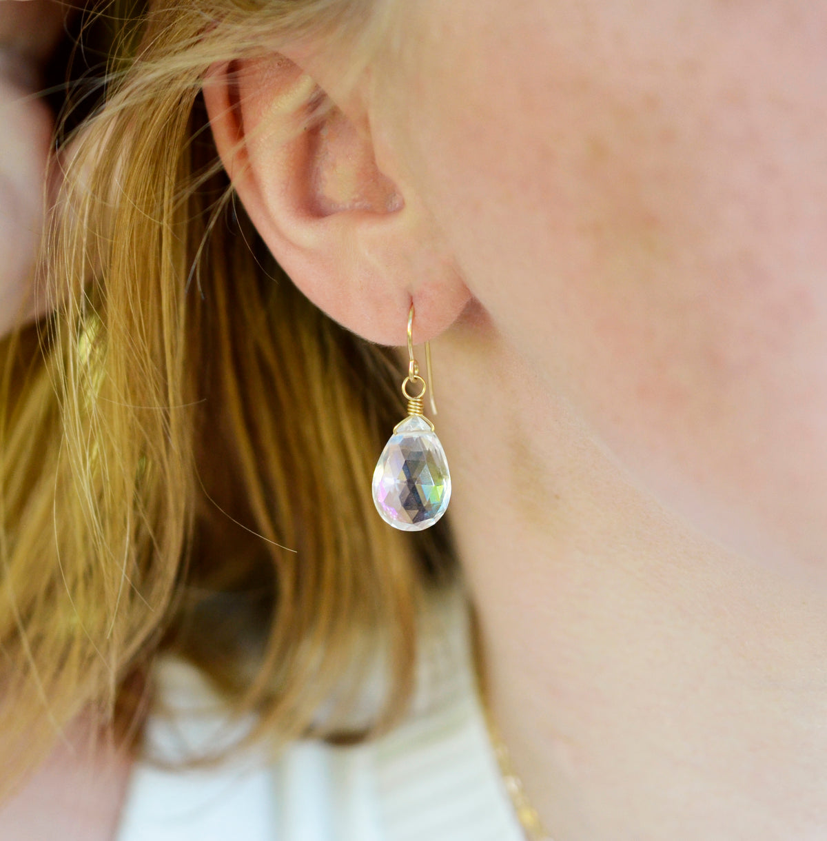 A woman wearing a rainbow mystic topaz faceted teardrop dangle earring in 14k gold filled. The crystals are clear, but contain rainbow colors that shift with movement. Handmade gemstone jewelry by Gemnia. 