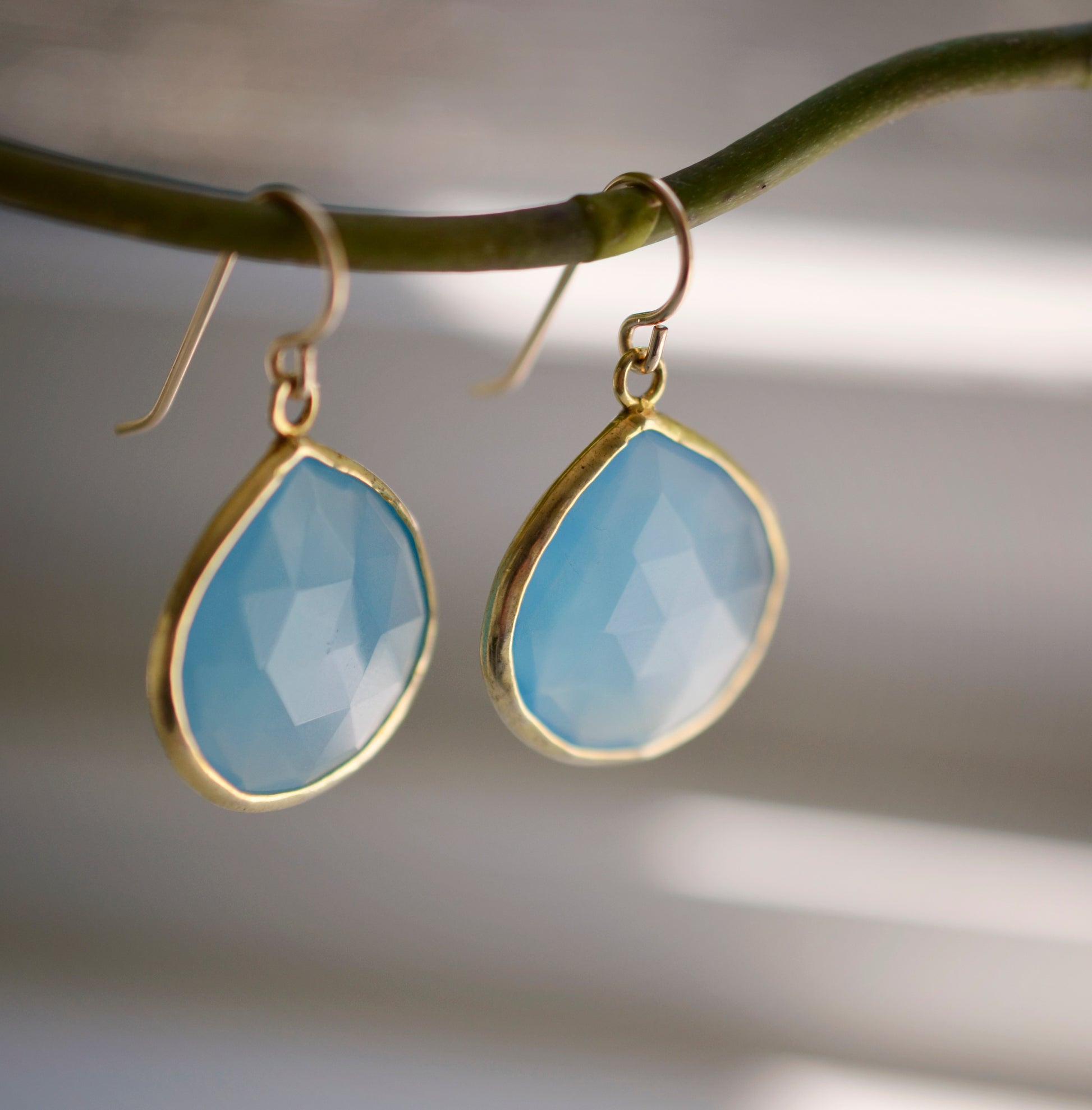 Large blue chalcedony teardrop gemstones bezeled in gold and suspended from earwires. Stones are faceted.