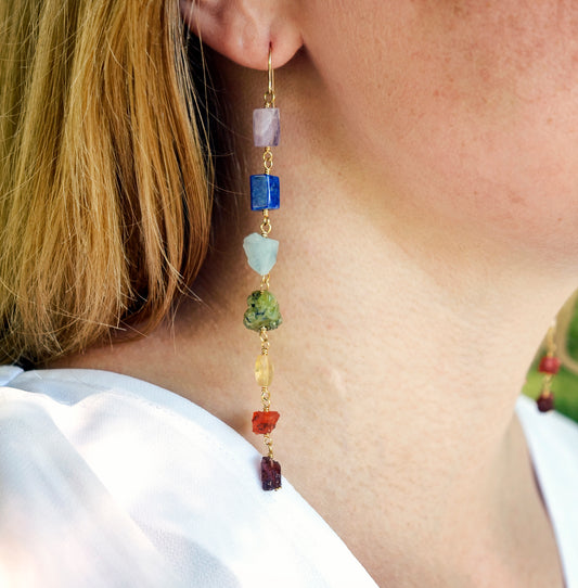 Long gemstone earrings made with a rainbow of raw gemstones. Colors include: purple, blue, aqua, green, yellow, orange, and red. Modeled image. 